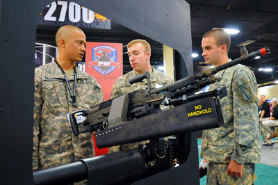 Three male soldiers stand around a rifle that is mounted on what appears to be a mockup of the side of a combat vehicle or helicopter.  A label on the rifle says "No handhold."  