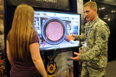 A woman and a man in a military uniform look at a television screen that appears to be part of a display at a conference.