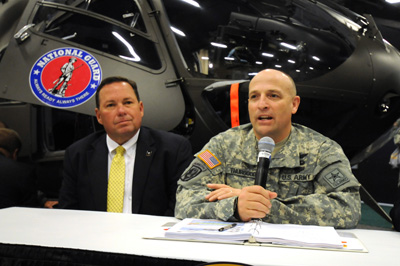 A man in a civilian suit and a man in a military uniform sit at a table.  The soldier has a microphone.  Behind them is a helicopter with a National Guard logo on the side.