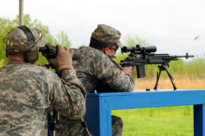 A soldier fires a rifle.  The copper shell casing flies off to the right.  Another soldiers sits behind him with a scope.