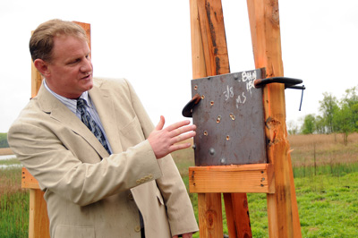 Outdoors, a man in a suit points at a sheet metal square mounted on a wooden rig. The metal square has been damaged by gun fire.