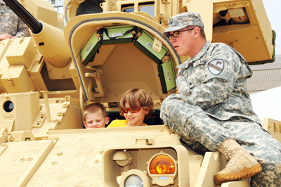 Two young boys peer out of an open hatch on a military vehicle. A soldier sits at the edge of the opening.
