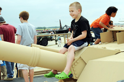A small, smiling boy sits atop a military tank and straddles the tank's gun barrel.  Another boy is nearby.  In the background other people are also standing on top of the tank.