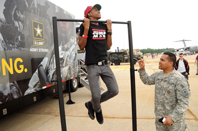 Outdoors, a young man in a red hat does pull-ups on a metal bar.  A man in a military uniform stands below and watches.  Behind them is a large vehicle with graphics on the side, including the U.S. Army logo.  In the far background is an airplane, people in a crowd, and other military vehicles.