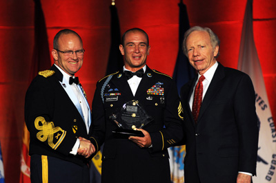 A man in a military uniform is holding an award. He is flanked by another man in a military uniform and a man in a civilian suit.  Behind them the wall is lit up in red lighting. Several flags are in the rear.