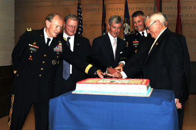 Six men, some in military uniforms and some in civilian suits cut a cake using a sword.  Behind them is an array of flags, and the "Pledge of Allegiance" is carved into the wall.