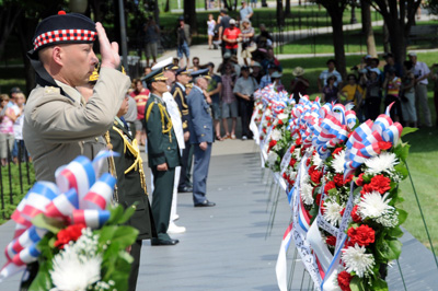 Outdoors, a row of men on the left, in military uniforms, stands before a row of floral wreaths, on the right.