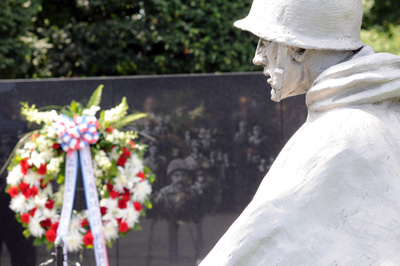 A statue of a soldier in a poncho and helmet is situated in front of a black slab wall with engravings on it.  A floral wreath sits in front of the wall.