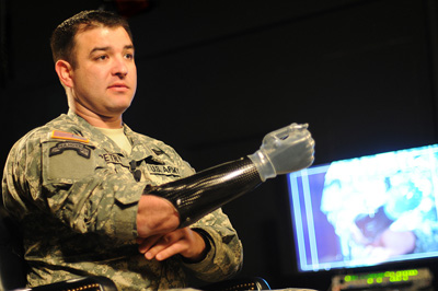 A man in a military uniform holds out his right arm, which is a prosthetic.  In the rear is a television screen. His uniform says "Petry" and "U.S. Army."