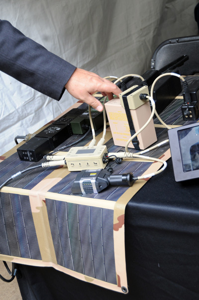 A hand touches electronic gear on a table. Several boxes and other devices are wired together and sit atop a flexible solar panel.