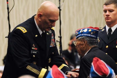A man in a military uniform interacts with an elderly man who wears a hat that indicates his status as a veteran. Another solider stands nearby.