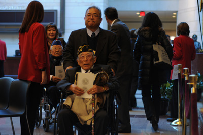 A man in a wheel chair wears a hat that indicates his status as a veteran.  Another man pushes him. Nearby is another man in a wheelchair.  