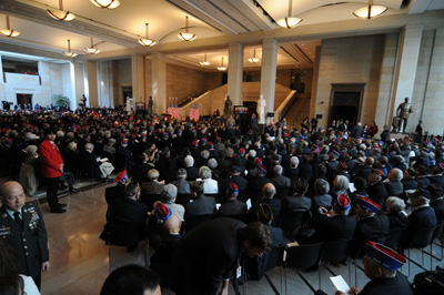 In a huge room with marble and pillars, hundreds of people are gathered most of them are sitting and facing towards the front of the room. American flags are on stands.  Multiple statues are in the room.
