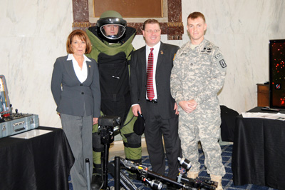A woman, a man in a protective suit for defusing bombs, a man in a suit, and a Soldier stand side by side.  In front of them is a bomb-defusing robot.  Nearby are other pieces of equipment.