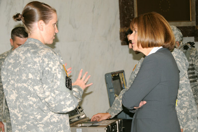 In a room with marble walls, a woman in a military uniform speaks with a woman in a suit.  In the background, Soldiers operate military equipment.