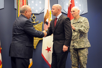 Two men in suits shake hands. Man in a military uniform stands nearby.  Behind them are multiple flags, including the U.S. Army flag.