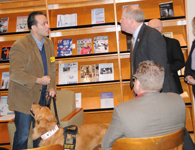 In a library, a man holds a service dog on a leash.  He speaks with another man.  Others are seated.