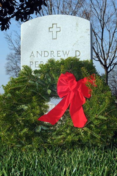 A wreath made of greenery and a red ribbon leans against a white gravestone. The gravestone says "Andrew D Perkins Jr."