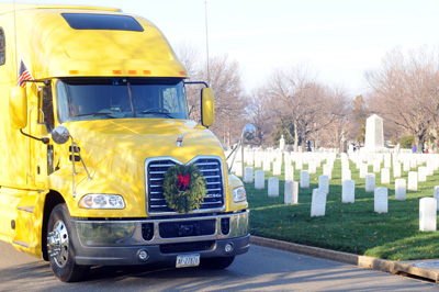 A large yellow truck is on a road inside a cemetery that has dozens of white gravestones.  A wreath is mounted t the front of the truck.