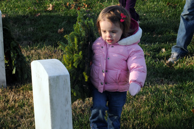 A tiny girl in  a pink coat approaches a white gravestone. She carries a wreath that is almost as big as she is.