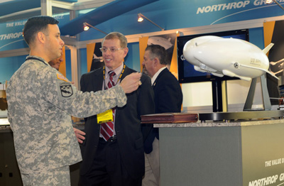 A man in a military uniform talks with a man in a suit. They stand next to a display of a tiny blimp.  The words "Northrup Grumman" are on the side of the pedestal that supports the blimp model. 
