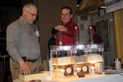 Two men stand behind a plexiglass apparatus that appears to contain three chambers filled with water and different types of silt.