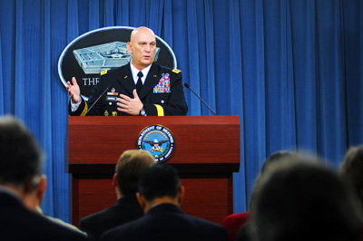 A man in a military uniform stands behind a lectern. Behind him is a sign with a picture of the Pentagon on it.  A crowd sits in front of him.
