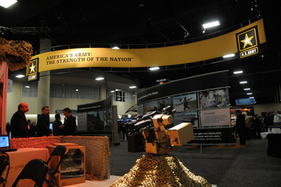 A robotic machine gun is situated in the middle of a large room with multiple military-related displays. A banner near the ceiling reads "America's Army: The Strength of the Nation."