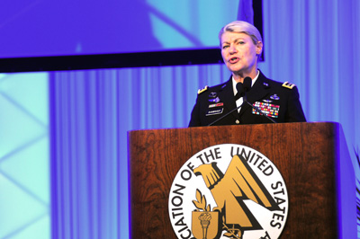 A woman in a military uniform stands behind a lectern. Behind her is a blue curtain. The lectern has a logo on it and the words "Association of the United States Army."