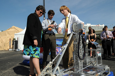 In an outdoor parking lot, an adult woman and a young man stand near a mechanical device that features a swinging robotic arm.  The young man points to the device.