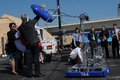 In a parking lot, a mechanical device's robotic arm has grasped the outstretched arm of an adult man.  A woman and several young adults stand nearby.