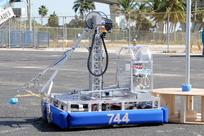 In a parking lot, a mechanical device with a swinging arm is mounted on a blue, mobile platform.  The number "744" is painted on the side.