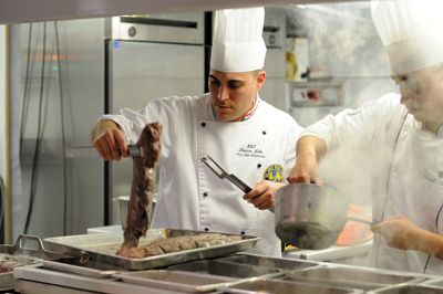 Two men in chef's outfits are in kitchen that is filled with steam. Once holds up a piece of meat.  The other is stirring something in a pan he is holding up. The entire kitchen is made of stainless steel.