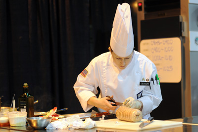 A woman in a chef's outfit cuts a string on a chunk of meat. 
