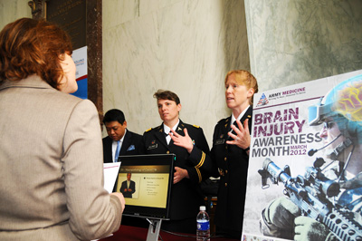 Two women in military uniforms stand behind a table with a man in a civilian suit.  A woman on the other side of the table talks with them. A sign nearby reads "Brain Injury Awareness Month March 2012."