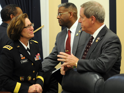 A woman in a military uniform talks to a man in a suit. Nearby, two other men shake hands.