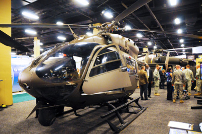 A helicopter sits indoors inside a convention center.  In the background, Soldiers and civilians are standing together talking.