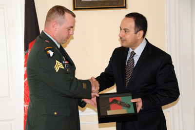 A man in a military uniform shakes hands with a man in a suit. The two both have their free hand on a frame with knife inside.