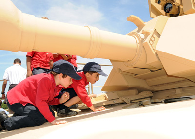 Two young boys climb on a tank. They are under the barrel of the tanks gun and peer through an open hatch.  
