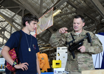 A young man in a blue shirt wears a dog tag around his neck on a chain.  He talks with a soldier who is handling a military rifle.