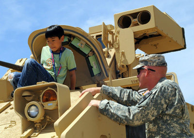 A boy crawls out the hatch on top of a military vehicle. a Soldier stands nearby.