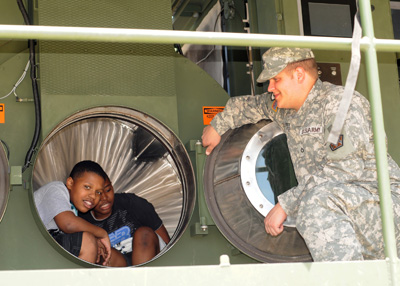Two boys are inside the drum of a large industrial clothes dyer.  A soldier sits outside and watches them.