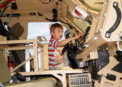 A young boy sits in the driver's seat of a large combat vehicle.  He presses bottoms on the dashboard.