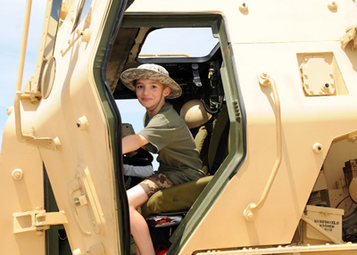 A young boy in a military-style hat sits in the driver's seat of a large combat vehicle.