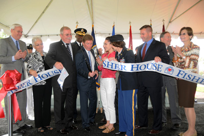 Nearly a dozen people stand together under a tent. One holds a pair of scissors and cuts a ribbon, which says "Fisher House."