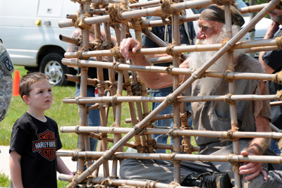 An elderly man with a long white beard is inside a cage made of bamboo.  A young boy stands outside the cage and looks at him.