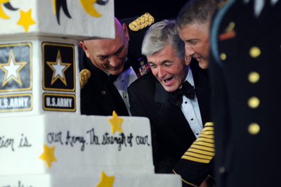 Three men, one civilian and two Soldiers,  use a sword to cut a large cake that says "U.S. Army" on it.
