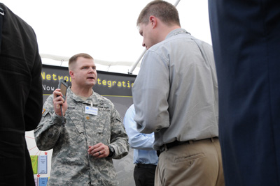 Under a tent, a civilian man talks with a soldier, who holds up an electronic device.