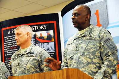 A man in a military uniform stands behind a lectern.  Next to him is another soldier. Behind the two is a wall hanging with a "Big Red One."