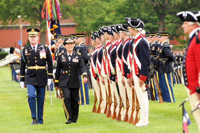 On a parade field, a man and a woman in military uniforms walk down a row of Soldiers who are dressed in Revolutionary War uniforms and who are standing at attention.